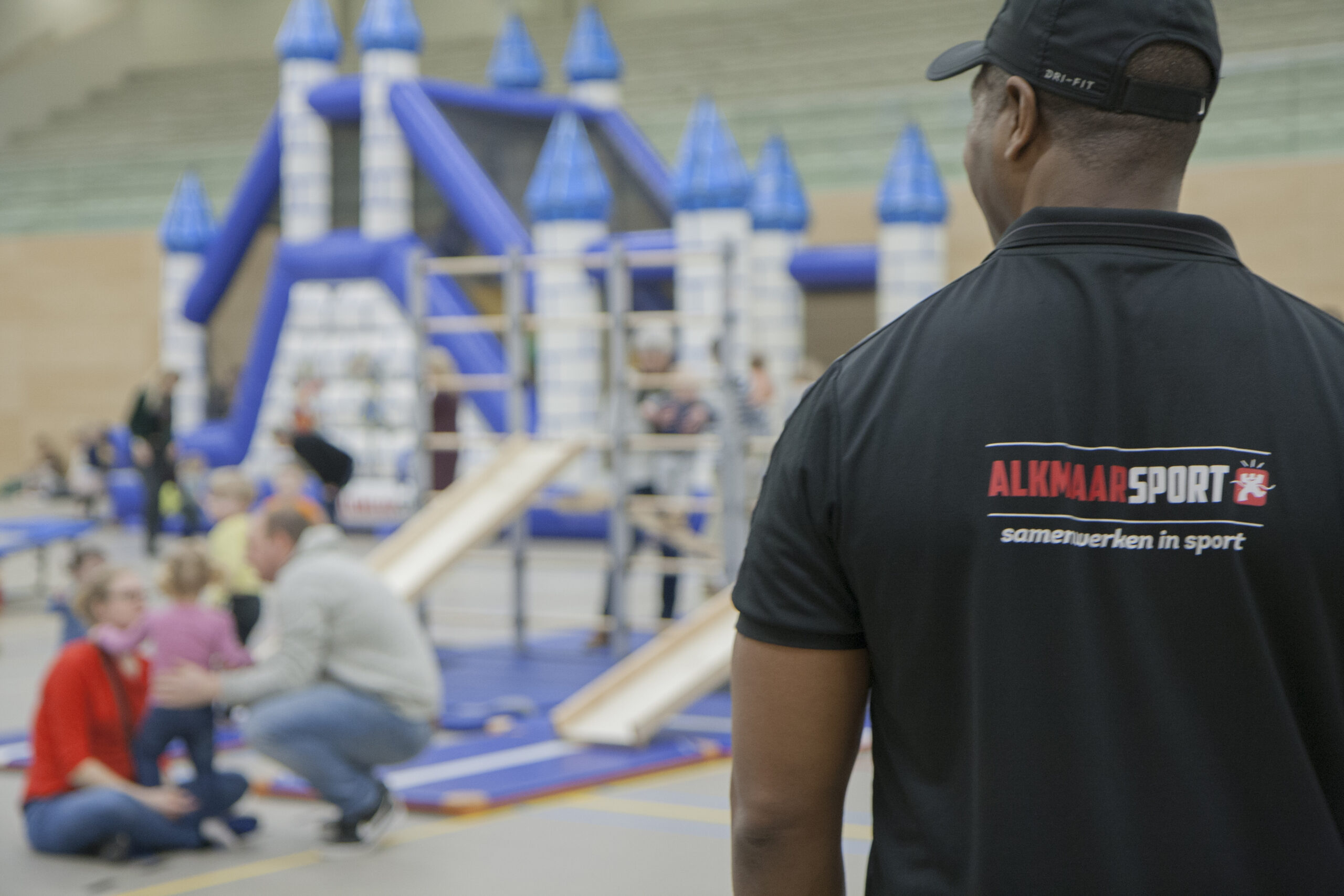 Kinderen in sportzaal, met de rug van trainer op de voorgrond die een t-shirt aanheeft van Alkmaar Sport