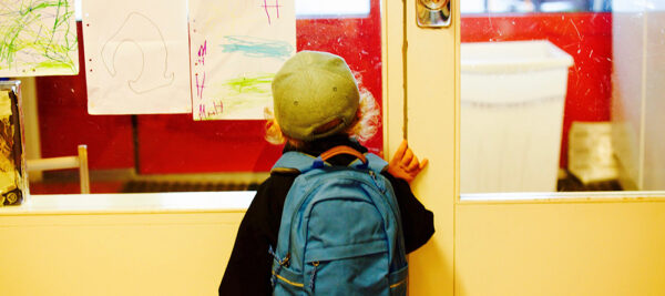 Foto van rug jongetje met rugzakje dat voor schooldeur staat.