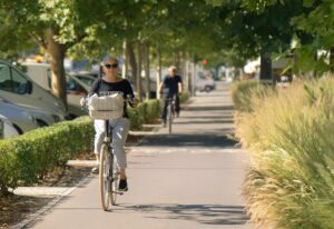 Foto van oudere man en vrouw op fiets in een straat met veel groen en geparkeerde auto's