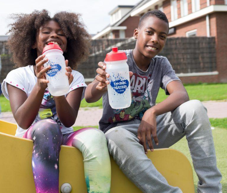 Een jongen en meisje in de tienerleeftijd buiten op een bankje en allebei een bidon water in de hand hebben
