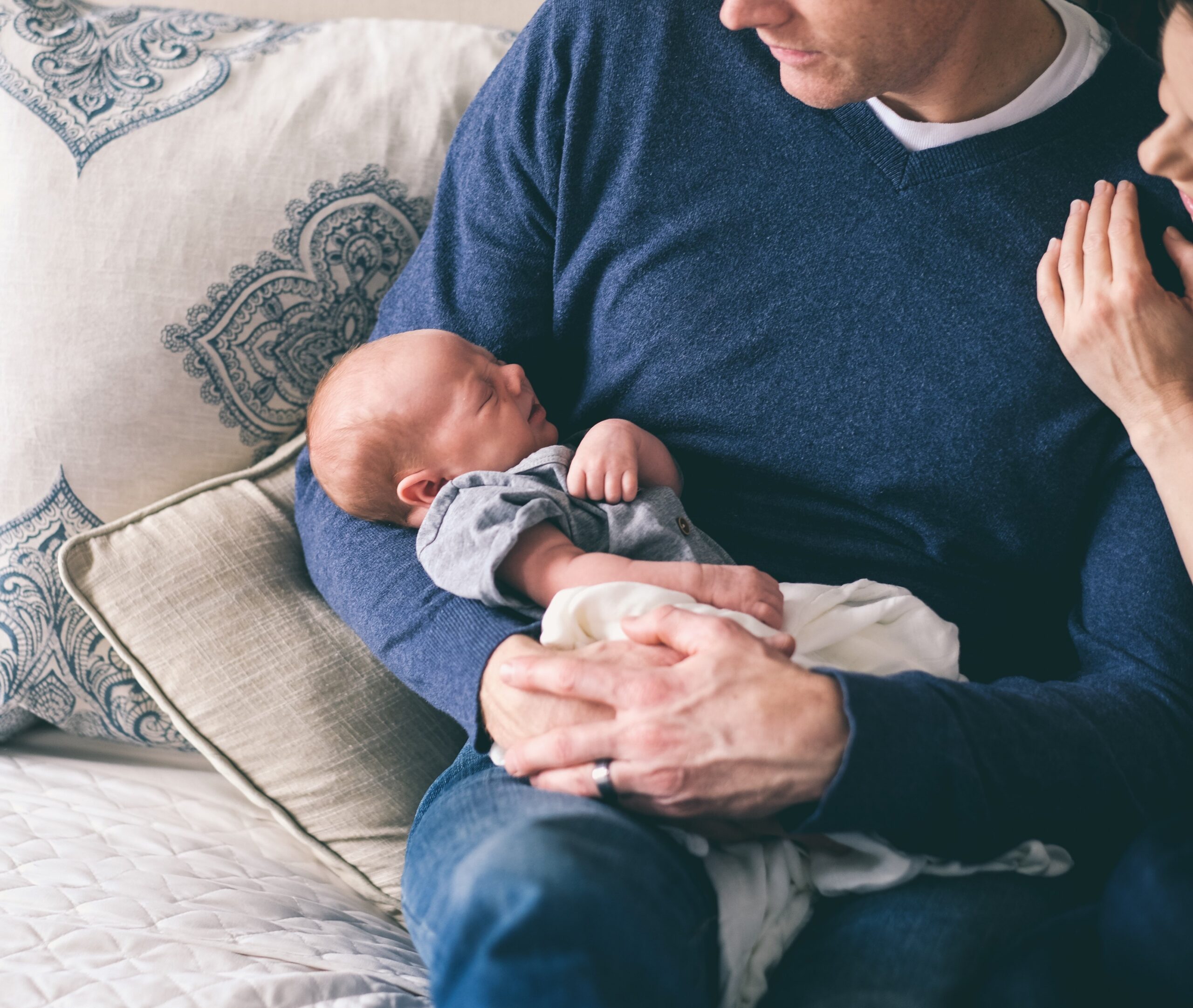 Vader in blauwe trui zittend op een bank met baby in zijn armen en nog net zichtbaar de hand van moeder op zijn schouder