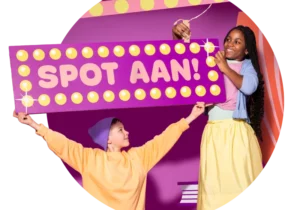 Ronde foto van twee kinderen tegen paarse theaterachtige achtergrond in de basisschoolleeftijd die een soort hardroze reklamebord omhoog houden waar SPOT AAN! op staat in hoofdletters