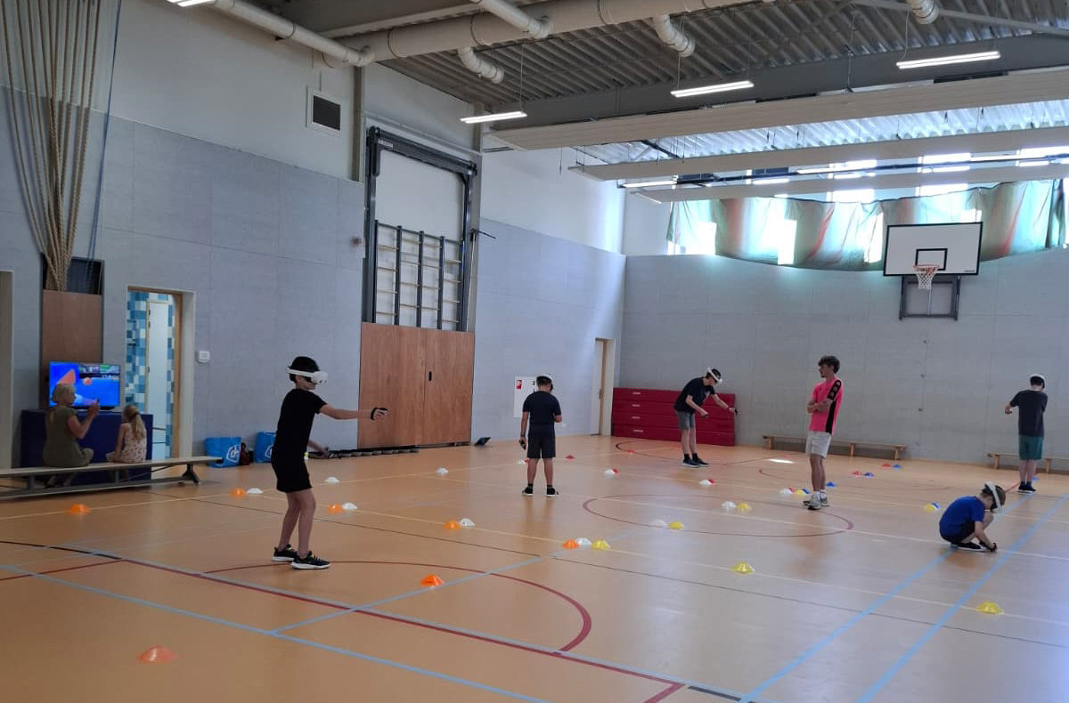 Foto van sportzaal met een paar kinderen in de basisschoolleeftijd en een begeleider tijdens een VR Fun, waarbij ze spellen spelen en bewegen met ene VR bril op